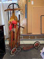 Vintage sled leaning against a wall beside a red hobby horse and a vintage metal scooter on a grey floor.