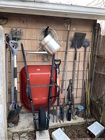 Full view of wheelbarrow frame with red plastic tray, watering can, various garden tools including shovels, pitchfork, hoe, digger, shears, stakes and bird feeders against outdoor wall.