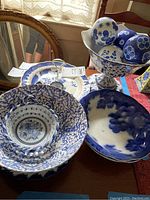 Various blue and white bowls, plate, and pedestal bowl with decorative balls displayed together on a table. Includes ceramic items with floral and nature motifs and lattice basket style bowl.