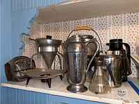 Shelf displaying assorted metal and glass beverage serveware including coffee and tea pots, carafes, dessert server