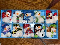 Top view showing 10 plush snowman ornaments assorted with different colored scarves and hats, resting in a blue box compartmentalized for each ornament.
