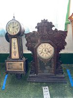 Full view of both clocks side by side on green carpet with lace curtains in background showing size and style differences.