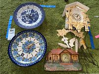 Four vintage clocks on green carpet including two decorative plate clocks, a bungalow clock, and a detached cuckoo clock with weights and pendulum.