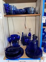 Shelf display showing multiple cobalt blue glassware items - plates, various glasses, parfait glasses, bottles, canisters, candle stick holder, and decanters.
