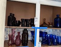 Shelves show various cranberry and amber glass items including cranberry glass bottles, glasses, creamer, sugar bowl, pitcher, and amber glass Krome-Kraft decanter, pitcher, and tray.