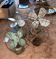 Four vintage metal table fans grouped together on carpet, showing visible rust and oxidation, various blade colors and styles.