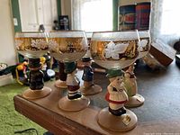Six vintage figural goblets arranged on a table displaying hand-painted glass tops with gold and white floral decorations and colorful ceramic figurine stems.