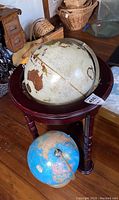 Photo showing two globes, a larger cream and brown toned world globe mounted in a dark wooden stand, with a smaller blue ocean floor globe below it on the floor.