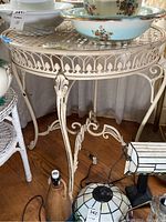 Full view showing the cream-colored round metal table with lattice top, decorative leaf trim, and curved legs. Scratches and worn paint visible.