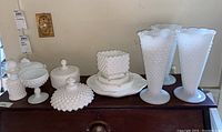 White hobnail milk glass collection displayed on a wooden dresser, showing two tall vases, a square candy dish, covered dishes with lids, and pedestal bowls.