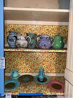 Shelving unit holding an assortment of vintage pottery pitchers and decorative plates and teapots. Pitchers display floral and multi-color painted designs. Plates and teapots are pastel shades with intricate patterns.