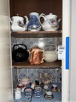 Full view of cabinet showing multiple shelves containing different teapots, tea cups, saucers, and small bowls arranged together. Variety of materials and colors.