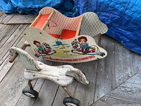 Photo of a white Radio Flyer wooden horse on wheels and a Raggedy Ann and Andy decorated wooden rocking horse seat on a porch, showing the front and side views with weathered paint and wear.