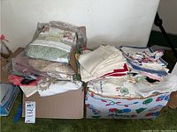 Two boxes filled with folded vintage linens including tablecloths, placemats, and napkins, shown on green carpet.
