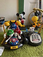 Two novelty push button phones on green surface, M&M themed phone with multiple colorful M&M characters in front, yellow handset on left side; Mickey Mouse figure in background