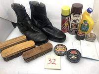 Pair of black leather boots with zipper and buckle, three wooden brushes, shoe polish tins, and leather care spray cans arranged together on white background.