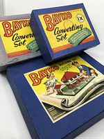 Three Bayko boxes: one Building Set in large blue box with colorful illustration of kids building house, two Converting Set boxes with yellow labels. All vintage 1950s style.