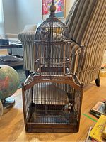 Front view of the antique bird cage showing wood frame, metal spindles, and fake birds inside. Background includes living room furniture and globe.