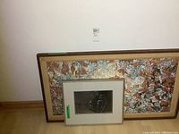 Photo of two framed artworks stacked against a wall. The smaller framed watercolor painting is placed in front of a larger colorful abstract framed piece.