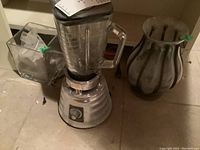Photo showing Oster classic blender, square glass vase, and rounded glass vase with metal accents on floor