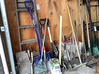 Garden tools arranged against a wooden wall showing various shovels, rakes, a bag of grass seed, and a ladder.