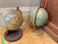 Two vintage-style globes on stands placed on a wood floor next to a wooden cabinet.