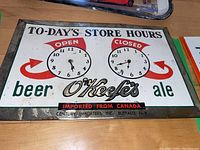 Full front view of vintage metal store hours sign showing O'Keefe's beer and ale branding, clock faces for open and closed hours, framed in metal with visible wear and rust.