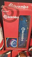 Photo showing a red boxed Brembo caliper cover in factory packaging marked HS-0313 with blue caliper cover visible inside.