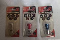 Three cigarette lighters in original blister packs with Noble R branding, showing clear, red, and blue tops.