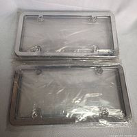 Two clear plastic license plate covers sealed in clear plastic wrapping lying flat on a white fabric background.