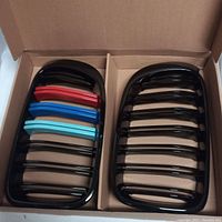 Pair of black glossy front grills with colored accents (red, blue shades) packed in a cardboard box