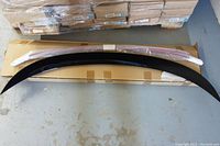 Photo showing black curved car spoiler lying on top of cardboard packaging on concrete floor with pallet stacked behind.