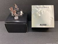 Bracelet shown upright on black stand next to original box with embossed classical head and Alessi logo