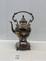 Front view of silver plated tilting teapot showing ornate handle and finial lid on decorative stand.
