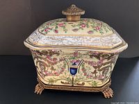 Front side view of the Asian covered dish placed on a brass footed stand, showing floral and gold detail with blue vase motif and brass finial on lid.
