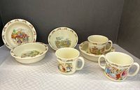 Photo showing set of 3 Royal Doulton Bunnykins cups and 4 bowls with colorful bunny illustrations on a white cloth.