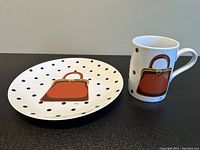 Photo showing full view of white ceramic cup and saucer, each decorated with a handbag design and black polka dots on a white background