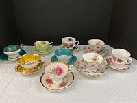 Nine tea cups with matching saucers displayed on white tablecloth in rows, showing floral and gold detailed patterns.