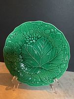Front view of an emerald green Majolica plate with embossed grape leaf and fruit design on display stand.
