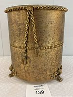 Front view showing the full gold metal waste paper basket with textured finish, rope molding and tassels at the top, and ornate claw feet base