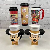 Front view of all five Disney plastic mugs arranged in two rows showing colors, designs, and shapes including travel mugs and Mickey Mouse head-shaped mugs.
