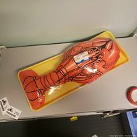 Photo of large stuffed lobster pillow shaped and colored like a lobster, on a yellow tray wrapped in clear plastic.