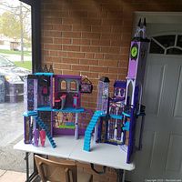 Front view of the purple and blue Monster High doll castle displayed on a white table outside, showing multiple levels, stairs, and small accessories.