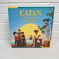 Front view of the Settlers of Catan Junior game box showing cover artwork with children, parrot, and sunset beach theme.
