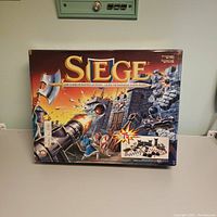 Front view of game box showing colorful siege battle artwork, game title SIEGE, and game piece count over 75 pieces.