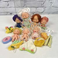 Seven vintage Strawberry Shortcake dolls sitting in two rows with assorted clothing and toys arranged in front of them on a white surface against a brick wall background.