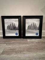 Two black 8x10 picture frames standing on floor against a wall, with placeholder photos of cypress trees.