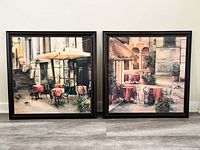 Pair of framed large square paintings showing outdoor cafe settings with tables, chairs, plants, and architectural features on stone plazas.