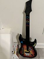 Black Guitar Hero wired guitar controller with colorful fret buttons and decals leaning vertically against a white wall next to four Nintendo Wii game cases.