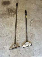 Two metal floor scrapers shown standing on floor, one longer and adjustable, the other shorter and fixed length, both with scraping blades attached.